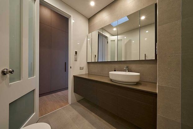 Contemporary bathroom with a long dark timber-toned floating vanity, white vessel basin, wall-to-wall mirrored cabinetry, and large taupe-grey tiled walls and floor. A skylight and recessed ceiling lighting provide balanced illumination, while a partially visible toilet and adjacent doorway confirm the bathroom layout.