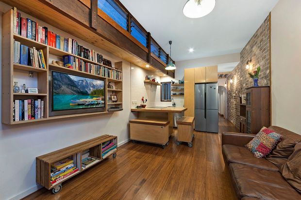 Compact open-plan living and kitchen interior with warm timber flooring, exposed brick walls, built-in timber shelving and media joinery, a leather sofa, and a raised mezzanine-level library with dark timber and blue-accent balustrade panels. The kitchen sits at the rear with timber cabinetry, a central island, stainless-steel refrigerator, and pendant lighting.