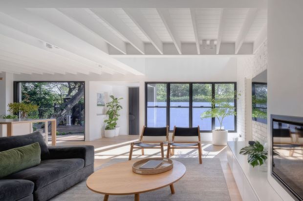 Bright, airy living room with white painted walls and exposed ceiling beams, large black-framed windows overlooking greenery and water, a dark charcoal sofa, two timber-framed lounge chairs, a round timber coffee table, indoor plants, and a built-in fireplace. The room has a relaxed contemporary coastal feel with abundant natural light.