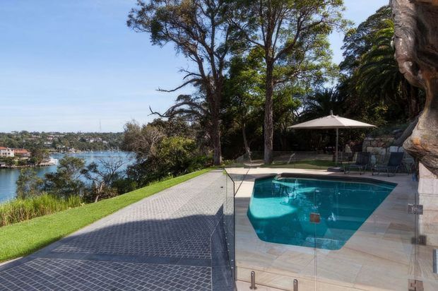A landscaped waterfront property with a turquoise swimming pool on a tiled terrace, bordered by clear glass pool fencing. A large dark grid-patterned paved area leads toward the pool and overlooks a calm inlet, while mature trees, a lawn, outdoor seating, and a large umbrella create a resort-like setting.