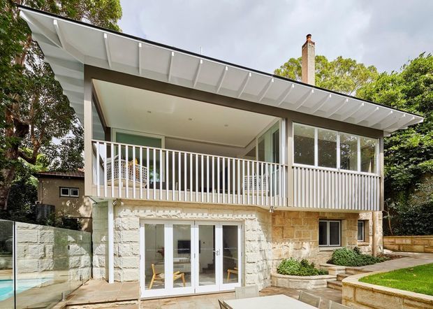 Exterior view of a two-storey residential house set among mature vegetation. The home has a projecting upper-level terrace with white vertical balustrades, extensive glazing, a broad shallow roof with exposed white eaves and fins, and a lower level faced in pale textured stone masonry. A small landscaped lawn, paved terrace, retaining walls and a partially visible swimming pool sit in the foreground.
