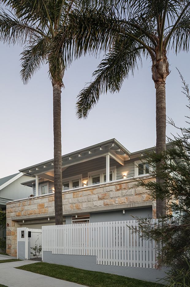 Street-facing view of a contemporary two-storey residence with a low-pitched roof, covered upper-level terrace, white posts and slatted screening, grey cladding, and a prominent band of light natural stone masonry. A white vertical-slat gate and boundary wall occupy the foreground, with tall palm trees framing the facade.