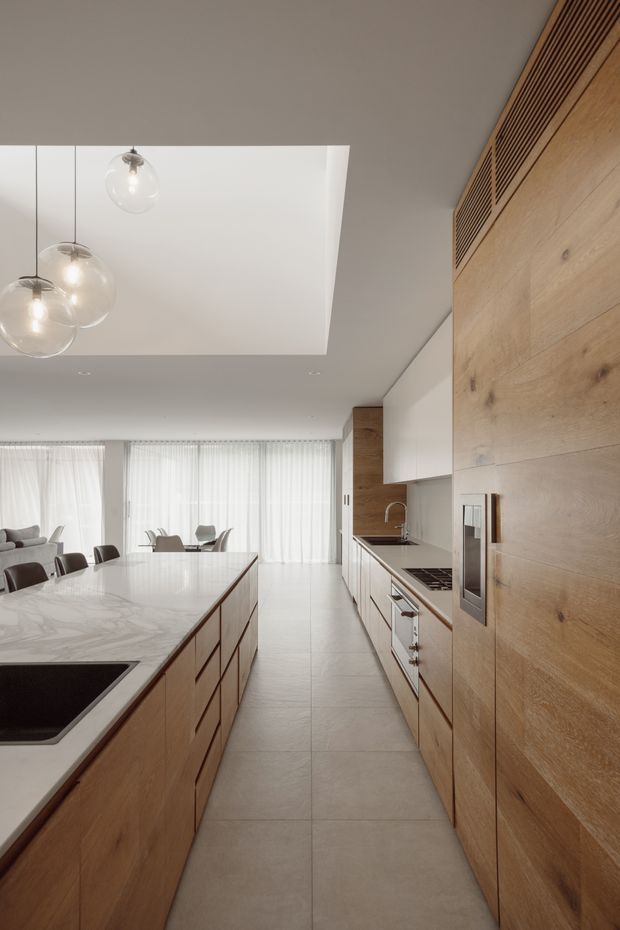 A bright, contemporary open-plan kitchen viewed along a central circulation aisle. The space features extensive light timber cabinetry and wall panelling, a large pale veined stone island with an integrated dark cooktop, matching stone benchtops, white overhead cabinets, full-height windows with sheer curtains, and three suspended clear glass globe pendants. A dining and living area is visible beyond the kitchen. The composition is clean, warm, and refined, with a large recessed ceiling feature above the island.