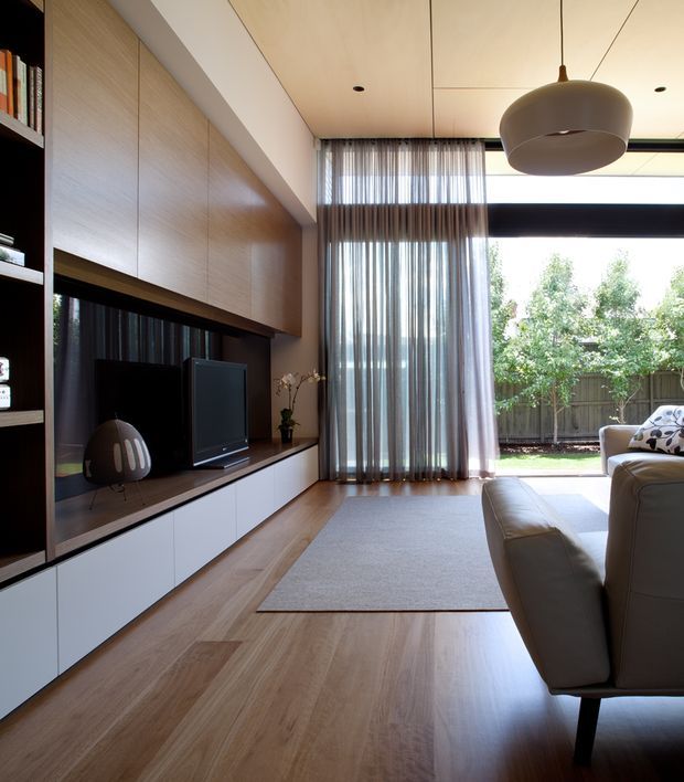 Contemporary living room with a long wall of integrated timber and dark-toned entertainment joinery, pale timber flooring, and a large neutral rug. Floor-to-ceiling sheer curtains frame generous glazing and a leafy outdoor view, while a sculptural pendant hangs from the light timber-lined ceiling. A sectional sofa occupies the foreground, creating a bright, refined interior with strong indoor–outdoor connection.