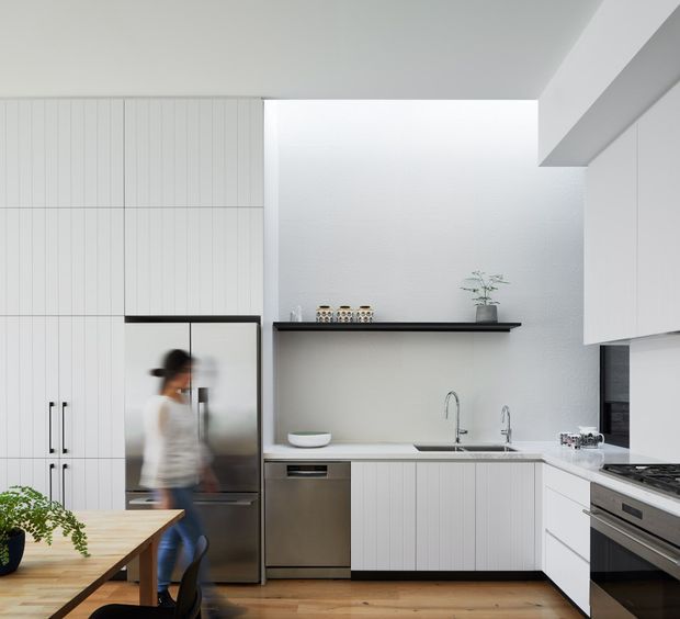 Bright contemporary kitchen with white vertically panelled cabinetry, a dark floating shelf, pale benchtops, stainless-steel appliances, timber flooring, and a timber dining table. A blurred person is present near the refrigerator, while a small plant and decorative items sit on the shelf.
