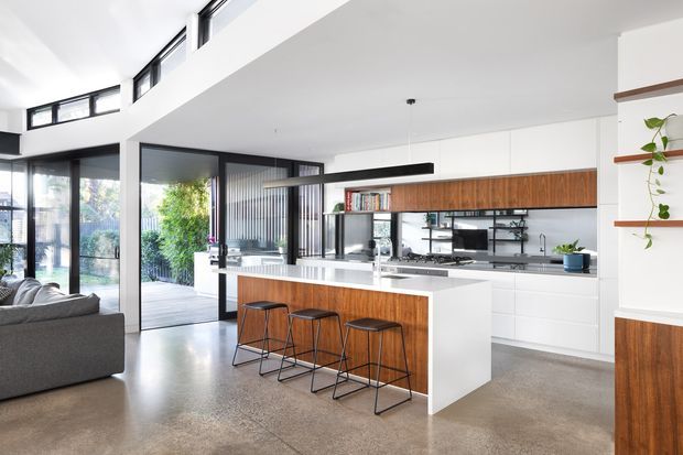 Bright open-plan kitchen and living area with a large white island featuring a timber-clad breakfast bar and three black metal stools. The kitchen has white flat-panel cabinetry, warm timber overhead joinery, open shelving, integrated appliances, and a long linear pendant light. Polished concrete flooring, expansive black-framed glazing, and a partially visible grey sofa contribute to a clean contemporary aesthetic.
