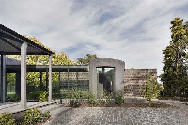 A contemporary residential exterior with a low-pitched dark roof, extensive floor-to-ceiling glazing, and a distinctive curved raw-concrete entry volume. The facade combines dark metal framing, textured brickwork, concrete steps, and a broad grey cobblestone-paved forecourt, with mature planting around the edges.