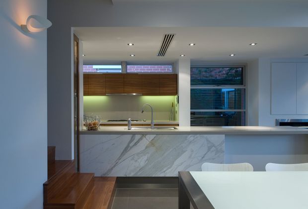 A contemporary open-plan kitchen and dining area viewed from a slightly raised timber-floored level. The kitchen features a large white marble-look island with integrated sink, pale benchtops, warm timber cabinetry, a green illuminated splashback, recessed ceiling lighting, and dark-framed windows. A dining table occupies the foreground, with stepped timber flooring and a wall sconce at left.