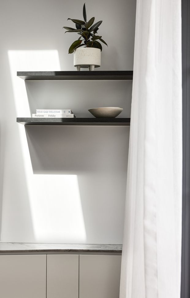 A softly lit minimalist interior vignette showing two dark floating shelves mounted on a pale wall, styled with a potted rubber plant, books, and a shallow ceramic bowl. Below is a low bank of pale built-in cabinetry with a dark stone-like or timber-look benchtop. A sheer white curtain partially frames the right side, while strong sunlight creates geometric shadows across the wall.