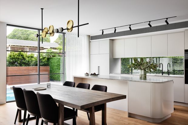Contemporary open-plan kitchen and dining area with a large dark timber dining table, black upholstered dining chairs, a white island with rounded ends, and extensive white built-in cabinetry. A mirrored splashback reflects greenery and cabinetry, while full-height glazing opens toward a landscaped outdoor area. Black track lighting and a sculptural brass-and-black pendant add contrast.