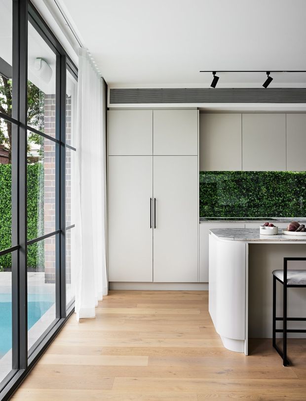 A bright, contemporary kitchen with full-height black-framed glazing along one side, overlooking a swimming pool and landscaped greenery. The space features pale flat-panel cabinetry, integrated tall storage, a curved white island with a light stone benchtop, timber flooring, sheer curtains, and a linear black track light. A compact black-framed bar stool sits at the island.