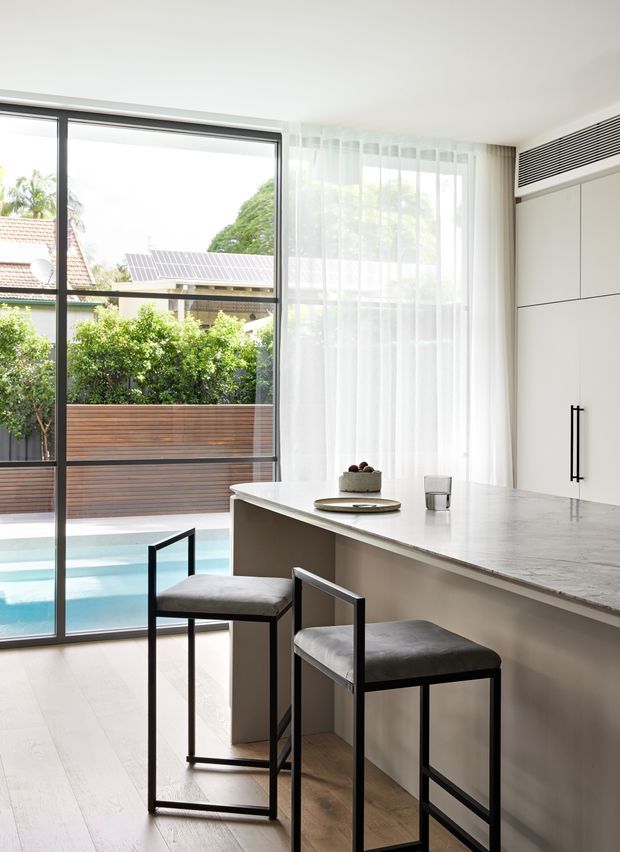Bright contemporary kitchen with a long pale stone island and two black-framed upholstered bar stools. Floor-to-ceiling glazing and sheer curtains open to a landscaped courtyard and swimming pool, while integrated white cabinetry and a linear air-conditioning grille create a clean, minimal interior.