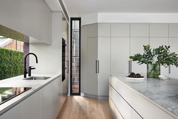 Bright contemporary kitchen with a long central island, pale integrated cabinetry, white and grey marble-look benchtops, timber flooring, and a black gooseneck tap. Full-height glazing and a narrow glazed door bring in natural light, with greenery visible outside and understated botanical styling on the island.
