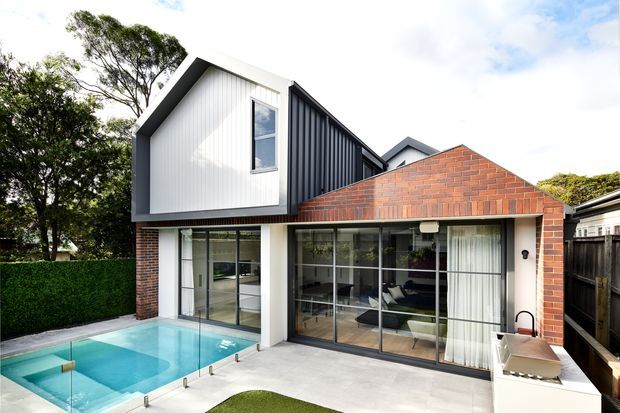 Contemporary two-storey residential extension viewed from a backyard pool area. The house combines white vertical cladding, dark standing-seam metal cladding, exposed red brick walls, extensive black-framed glazing and a steep gabled roof. A paved terrace, swimming pool and built-in outdoor cooking area occupy the foreground.