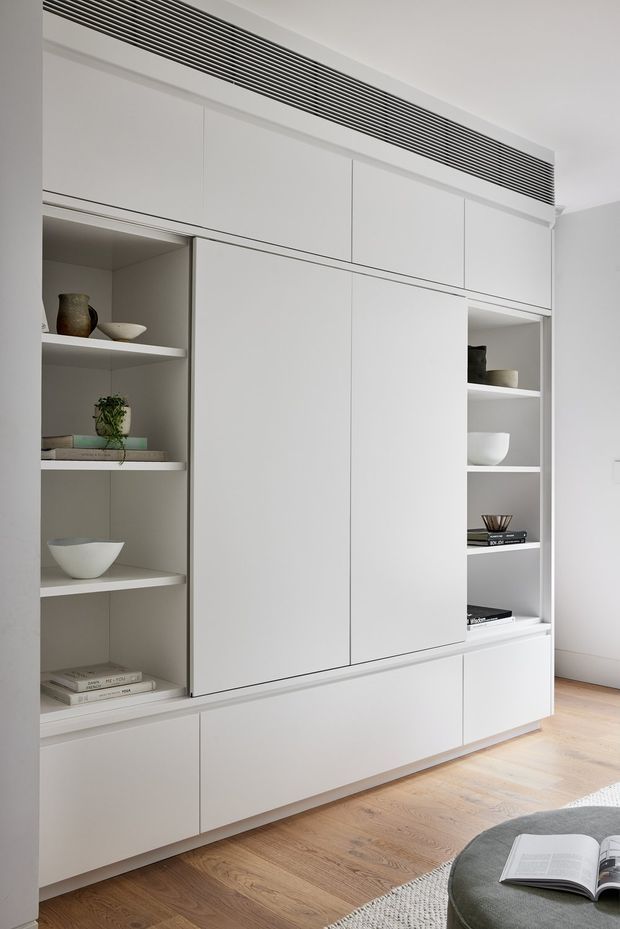 A bright, minimalist built-in storage wall featuring extensive matte white cabinetry, integrated open display shelving, and a long linear ventilation grille near the ceiling. The joinery is set against pale walls and a light timber floor, with neutral decorative objects and part of a rounded lounge chair visible in the foreground.
