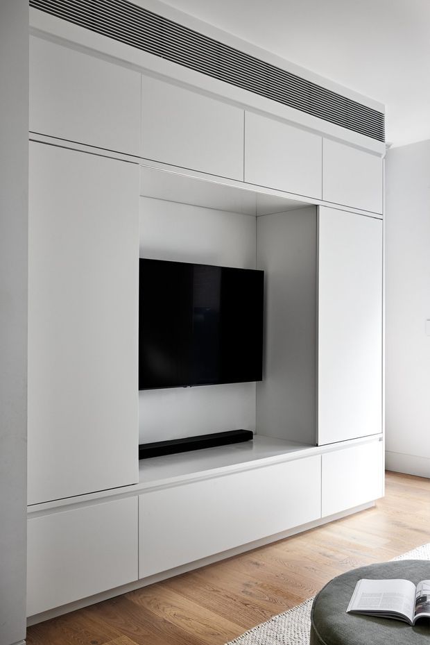 A minimalist built-in entertainment wall with floor-to-ceiling white cabinetry, concealed storage, a recessed niche containing a wall-mounted black television and slim media shelf, and a linear black ventilation grille above. Pale timber flooring and the edge of a neutral rug and upholstered seating are visible in the foreground. The space is bright, clean, and highly streamlined.