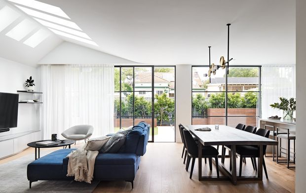 Bright open-plan living and dining area with a deep blue sectional sofa, black dining table and chairs, and full-height glazing opening to a leafy suburban outlook. A large angled skylight brings daylight into the white interior, while slim black-framed windows, pale timber flooring, and minimal built-in shelving create a clean contemporary feel.