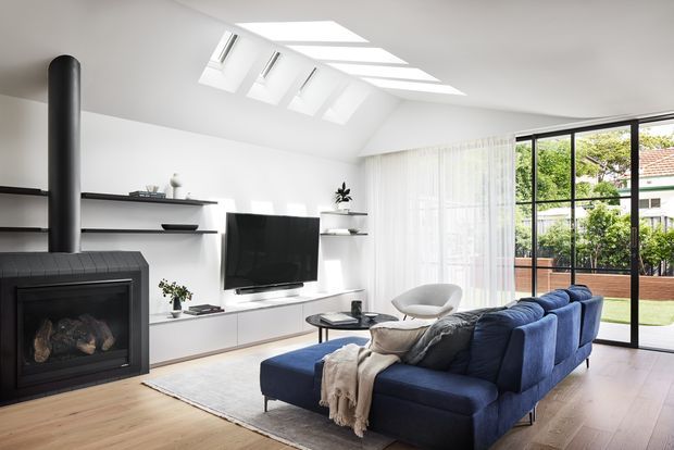 Bright contemporary living room with a deep blue sectional sofa, black wood-burning fireplace, minimalist white built-in media cabinetry and open shelving, pale timber flooring, and expansive black-framed glazing opening to a courtyard. Multiple roof skylights and sheer curtains provide abundant natural light.