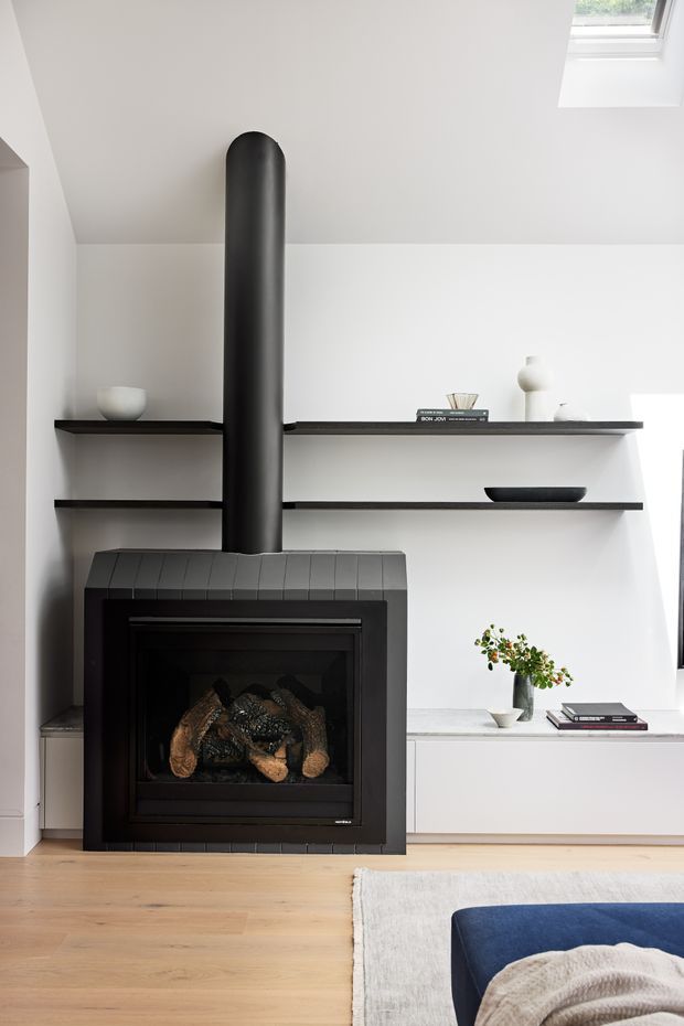 A bright, minimalist living room featuring a black freestanding wood-burning fireplace with a tall cylindrical flue, integrated white built-in shelving and cabinetry, pale timber flooring, and restrained black-and-white decor. A skylight brings natural light into the room, while a blue upholstered sofa and rug are partially visible in the foreground.
