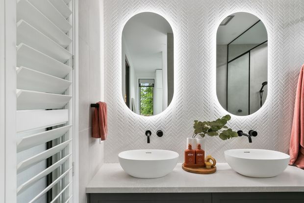 Bright contemporary bathroom with a double vanity, two white vessel basins, arched mirrors, a white herringbone-textured wall finish, dark cabinetry, and a large white louvred window or screen. A glass shower enclosure is visible in the mirror reflection, with coral towels and minimal styling adding warmth.
