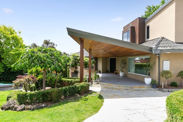 A bright exterior view of a contemporary residential entry and covered outdoor entertaining area. The home combines a projecting flat canopy with a timber-lined soffit and timber posts, a beige rendered facade, dark timber upper cladding, and an existing tiled pitched roof. A paved walkway leads through manicured garden beds toward the entry, with a swimming pool partially visible at the left.
