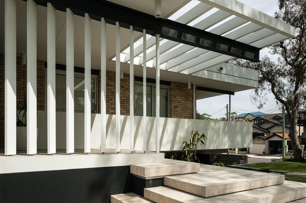 Exterior view of a contemporary multi-level residence with a strong geometric facade. White vertical fins screen the entry and glazing, contrasting with exposed tan brick walls and a black structural beam. Broad concrete steps lead to a raised front terrace beneath a translucent pergola or canopy, with neighbouring suburban homes and established trees visible beyond.