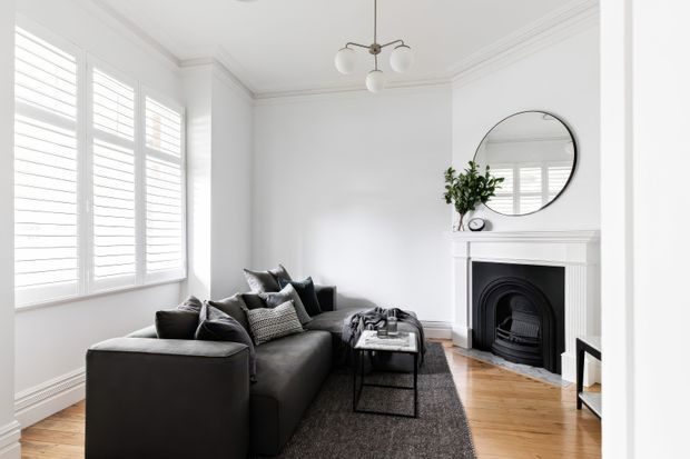 Bright, refined living room with a charcoal sectional sofa arranged around a small black coffee table and dark textured rug. White walls feature traditional cornices and a classic fireplace with mantel, while plantation shutters provide abundant filtered daylight. A round mirror, potted plant, and contemporary multi-globe pendant add restrained decorative detail. Timber flooring and monochromatic furnishings create a clean, elegant contrast.