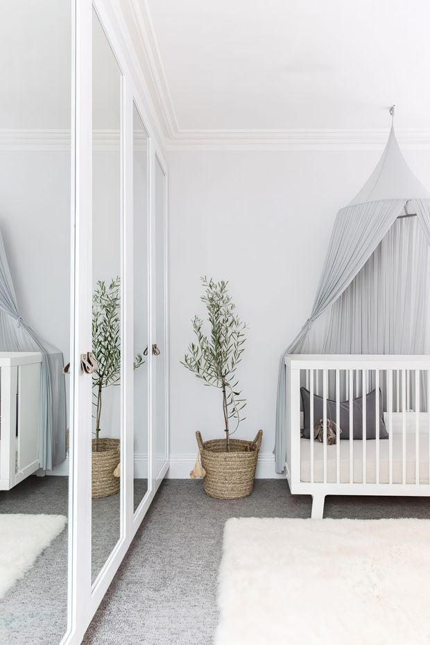 Bright, serene nursery with a white cot beneath a draped canopy, mirrored wardrobe doors, grey carpeting, white faux-fur rug, and potted olive trees in woven baskets. The room features crisp white walls with decorative ceiling cornices and a soft grey-and-white palette.