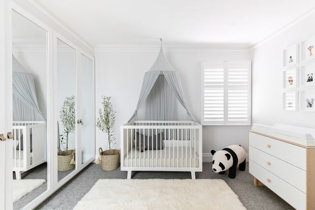 Bright, serene nursery with a white cot centered beneath a draped canopy, mirrored built-in wardrobe doors on the left, a white dresser on the right, plantation shutters, wall shelving, soft grey carpet, and a plush white rug. The room uses a restrained white and light-grey palette with natural woven baskets and gentle decorative details.