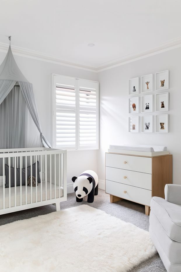 Bright, softly styled nursery with a white cot beneath a draped canopy, white plantation shutters, a white and timber chest of drawers with changing top, framed animal artwork, a plush panda toy, and a white armchair. The room has pale grey walls, white decorative ceiling moulding, grey carpet, and a large soft white rug, creating a clean and serene atmosphere.