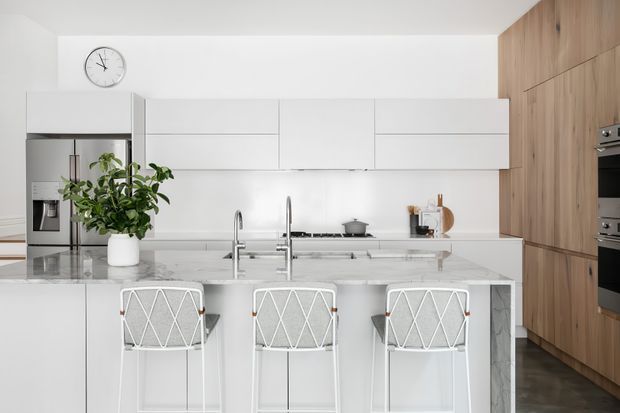 Bright contemporary kitchen with a large island and three bar stools. The space features white flat-panel cabinetry, a pale stone island benchtop, integrated stainless-steel appliances, timber-toned full-height cabinetry, dark flooring, and a large potted plant. A minimalist black wall clock and simple countertop accessories add restrained styling.