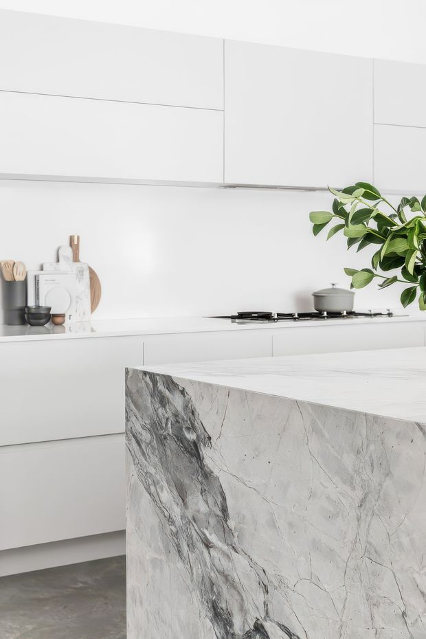 A bright, minimalist contemporary kitchen in predominantly white tones. The space features sleek handleless cabinetry, a long white benchtop with integrated cooking appliances, and a substantial waterfall island clad in dramatic white-and-grey veined natural stone. A few timber kitchen accessories and a leafy plant add warmth and contrast.