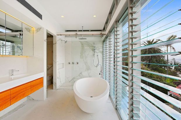 Bright contemporary bathroom with a freestanding white oval bathtub beside full-height louvred glazing. The room features pale marble-look wall and floor surfaces, a floating vanity with vivid orange timber cabinetry and a white benchtop, frameless glass shower enclosure, mirrored cabinetry, and exposed ceiling services. Tropical greenery and a palm are visible beyond the louvres.