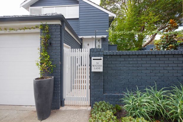 Exterior view of a residential property entry, showing a dark blue-grey painted brick boundary wall, white vertical slat pedestrian gate, white garage door, and a two-storey house with dark horizontal weatherboard cladding and a gabled roof behind. Established greenery and sculptural planting frame the entrance, with a visible address plaque reading 34 Waters Lane.