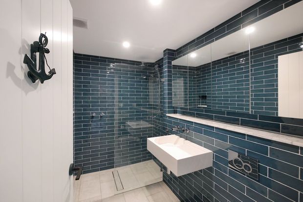 Contemporary bathroom featuring dark blue-green subway tile walls with contrasting light grout, a large mirrored vanity wall, floating rectangular white basin, wall-mounted fixtures, and a tiled walk-in shower. Light floor tiles and white cabinetry create contrast, while recessed ceiling lighting provides an evenly illuminated finish.