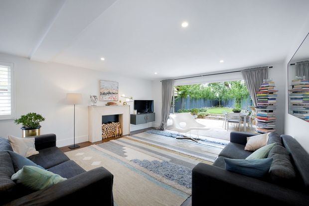 Bright contemporary living room with two dark sofas arranged around a white fireplace and wall-mounted television. A large patterned area rug spans the room, with a floor lamp, coffee table, cushions, and stacked firewood adding warmth. Full-height glazed sliding doors open to a furnished outdoor terrace and garden, while built-in shelving and a large mirror line the right wall.