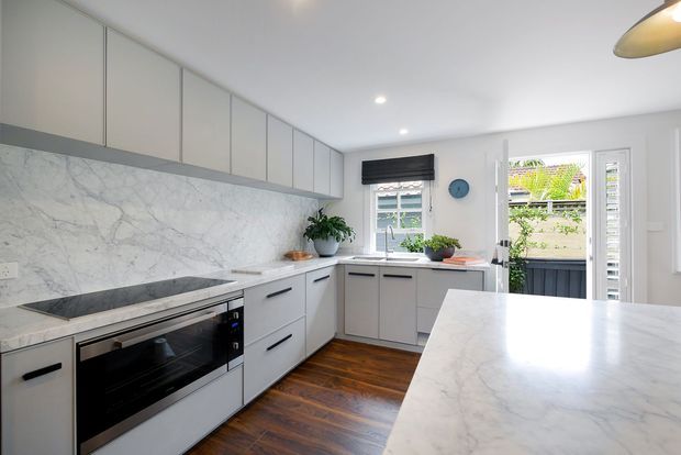 Bright contemporary kitchen with extensive pale cabinetry, a marble-look benchtop and splashback, integrated black oven and cooktop, timber flooring, and a large foreground island. White walls, recessed ceiling lights, indoor plants, and windows overlooking greenery create an airy residential interior.