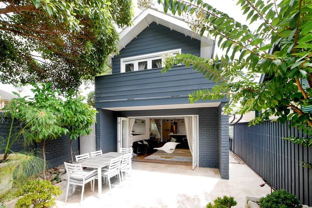 A compact two-storey residential extension or cottage viewed from a landscaped outdoor entertaining area. The house has dark blue-grey horizontal weatherboard cladding, crisp white trim, a pitched gable roof, and a projecting upper-level balcony enclosed by matching cladding. Large bi-fold or folding glazed doors open from the lower level onto a pale paved patio with white outdoor dining furniture. Dense greenery and dark fencing frame the facade.