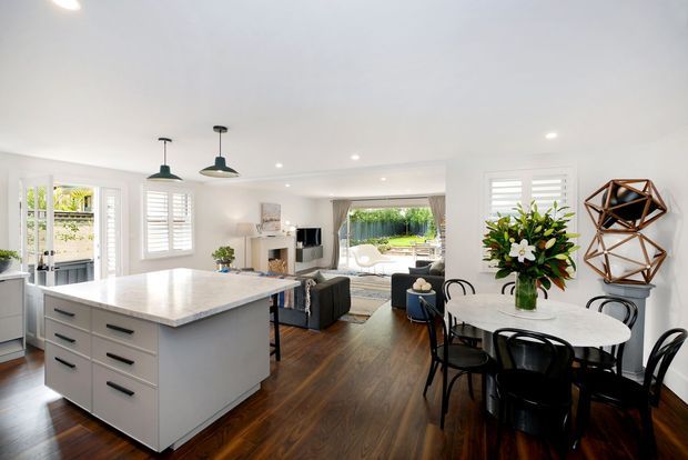 Bright open-plan kitchen and dining area flowing into a living room, with a large white stone island, white shaker-style cabinetry, dark timber flooring, pendant lights, plantation shutters, a round dining table, and views through the living area to an outdoor space. The interior is contemporary with crisp white finishes and dark contrasting furnishings.