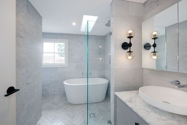 Bright contemporary bathroom featuring a freestanding white bathtub behind a frameless glass shower screen, a white vessel basin on a pale marble-look vanity, large light stone wall tiles, herringbone floor tiling, mirrored cabinetry, and distinctive black-and-brass globe wall lights. A window provides natural light.