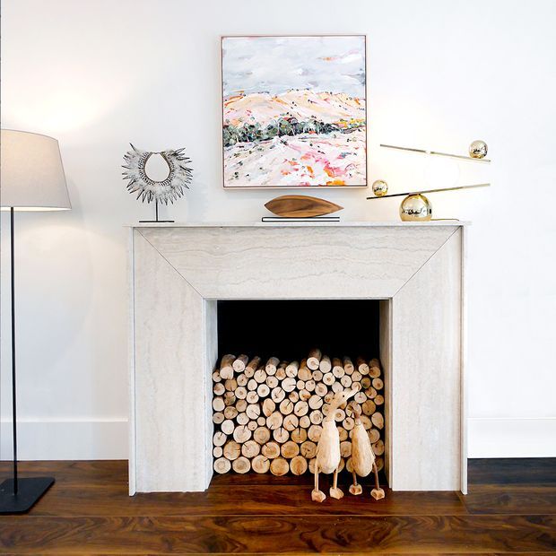 A refined living room vignette centered on a pale stone fireplace surround filled with stacked firewood. The white wall has simple panel moulding, while dark patterned timber flooring adds warmth. Mid-century-inspired brass artwork and sculptural decor sit on the mantel, with a large abstract landscape painting above and a floor lamp at left.