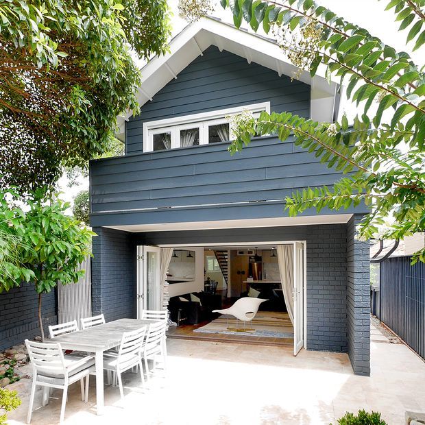 A compact two-storey residential extension or dwelling viewed from a leafy courtyard. The facade combines dark blue-grey horizontal weatherboard cladding on the upper level with dark painted brick around the ground-floor opening, white trim, and a steep gable roof with decorative eaves. Wide bi-fold or folding doors open onto a paved outdoor dining area, revealing a bright interior with a small dining or living space and staircase.