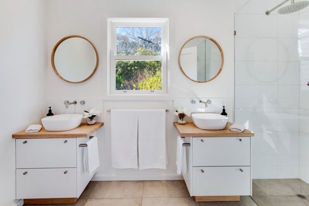 Bright, symmetrical contemporary bathroom with two white vessel basins on timber-topped white vanities, round timber-framed mirrors, a central window with white trim, white wall tiling, and a glass-enclosed shower with an overhead rain shower. The room has a light, clean, balanced appearance.