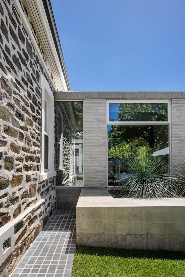 Exterior architectural view of a contemporary extension adjoining an older stone masonry building. The composition features a rough natural-stone wall, pale brick cladding, strong concrete lintels and planter walls, large black-framed glazed openings, and a tiled courtyard path with sculptural planting.