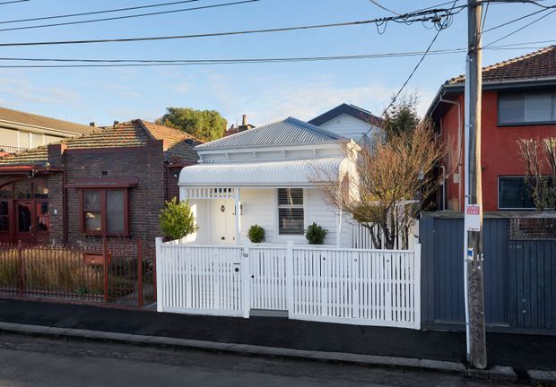 Street-facing view of a compact single-storey residential house in an established urban neighbourhood. The home has a white weatherboard facade, decorative verandah detailing, a corrugated metal gable roof, white picket-style fencing and gate, and a small planted front garden. Adjacent brick and rendered homes, overhead utility wires, and a timber utility pole are visible.