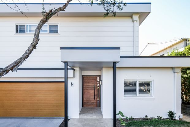 Front exterior of a contemporary two-storey residence with a flat roof, crisp white horizontal cladding, dark trim, a timber garage door, and a sheltered entry canopy supported by slender dark posts. The facade includes horizontal windows with louvres and a timber front door, with established landscaping in the foreground.