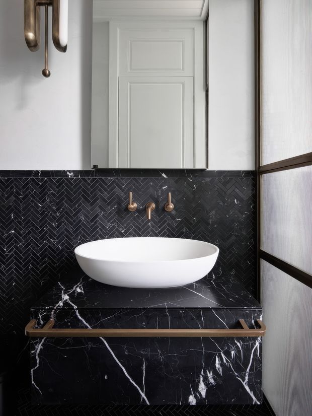 A compact, refined bathroom vanity featuring a white vessel basin set on a dramatic black marble counter and cabinet. The wall behind is clad in black marble mosaic tiles arranged in a herringbone pattern, with brushed brass wall-mounted tapware and a matching sculptural wall light. White paneled cabinetry and a textured translucent glass screen complete the high-contrast, luxurious composition.