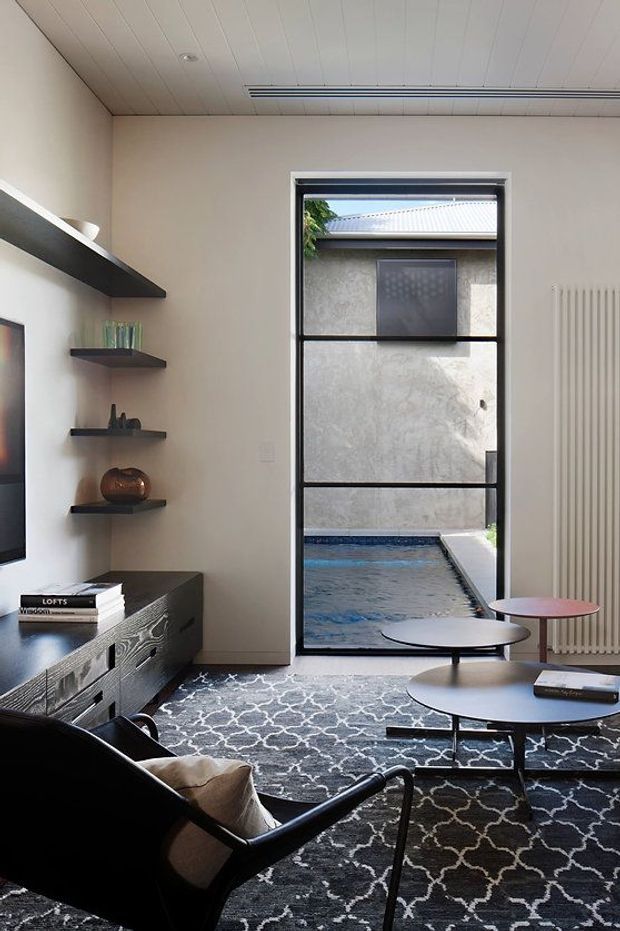 A refined contemporary living room with a black-framed glazed door opening directly to a small courtyard pool. The room features a dark timber media console, floating display shelves, patterned dark rug, sculptural black lounge chair, and layered round coffee tables. A pale vertical-panel ceiling, neutral walls, linear air-conditioning vent, and radiator create a clean architectural backdrop, while the pool and adjacent masonry wall are visible outside.
