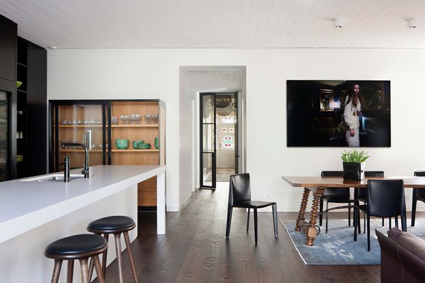 Bright contemporary open-plan kitchen and dining area with a long white island, integrated sink and black stools, adjoining timber-glass display cabinetry, dark timber flooring, and a dining table with black chairs. A glass-panelled doorway leads to another space, while a wall-mounted television and large rug sit in the dining zone. The white tongue-and-groove ceiling and restrained black, white, and timber palette create a clean, refined interior.