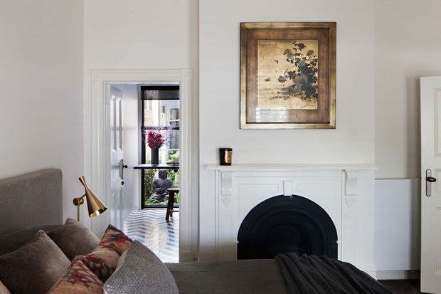 A refined living room with a grey upholstered sofa, patterned cushions, a brass reading lamp, and a traditional white-painted fireplace mantel with a dark arched firebox. White wall panelling and trim frame the room, while a framed landscape artwork hangs above the mantel. A glazed doorway and adjacent door provide views into adjoining spaces.